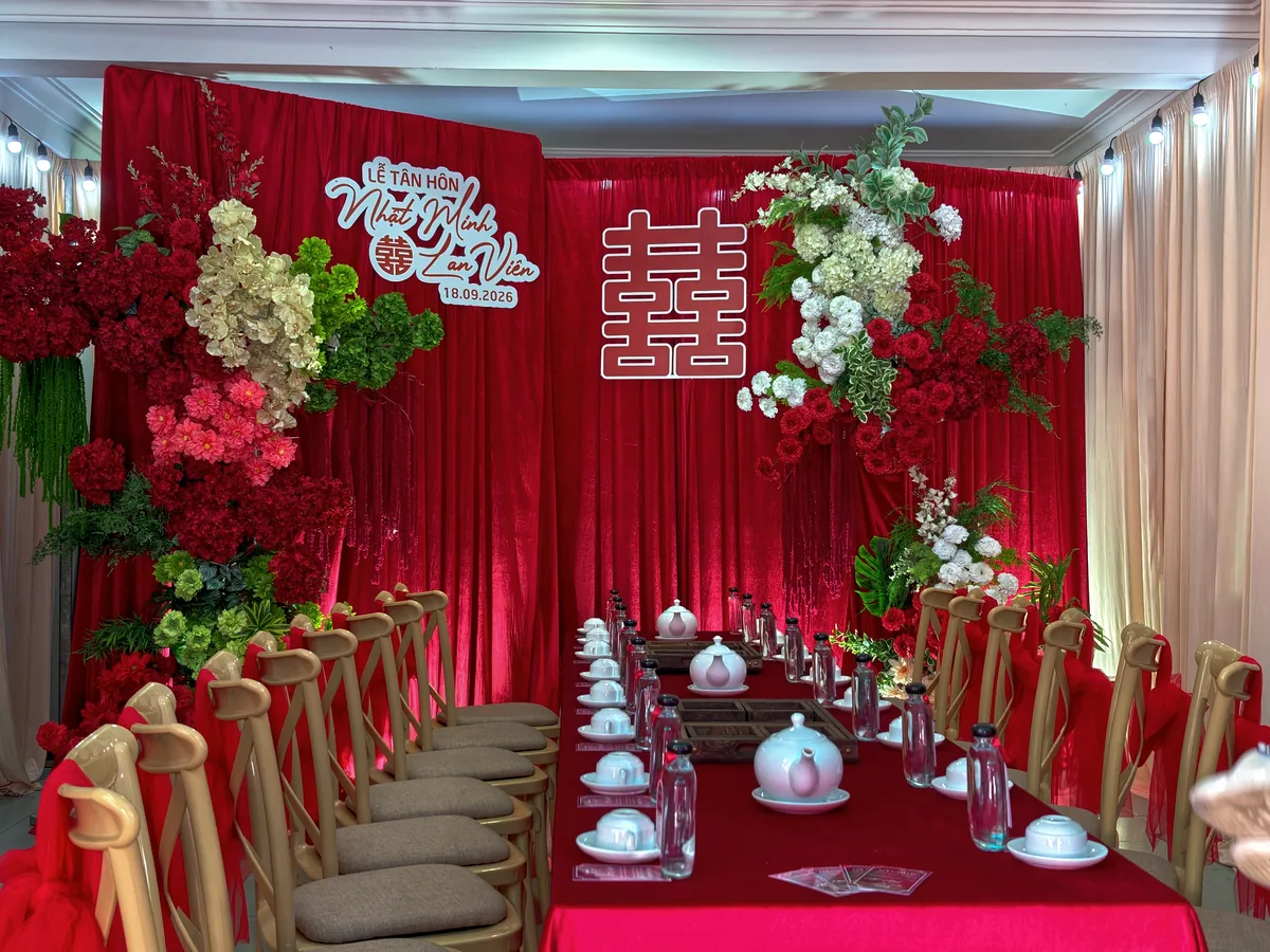 Không gian lễ tân hôn tông đỏ với backdrop chữ hỷ, hoa lụa và bàn tiệc dài phủ khăn đỏ